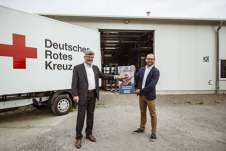 Rüdiger Unger, Vorstandsvorsitzender des DRK Landesverbandes Sachsen, und Alexander Hesse, Marketingleiter bei PostModern, halten gemeinsam die große Sonderbriefmarke fest. Im Hintergrund stehen die Logistikhalle und ein LKW mit dem roten Kreuz und der Aufschrift Deutsches Rotes Kreuz