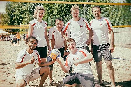 Sechs Volleyballspielerinnen und Volleyballspieler stehen auf einem Strandvolleyballfeld und lächeln in die Kamera. Zwei der Spieler halten den Volleyball in den Händen.