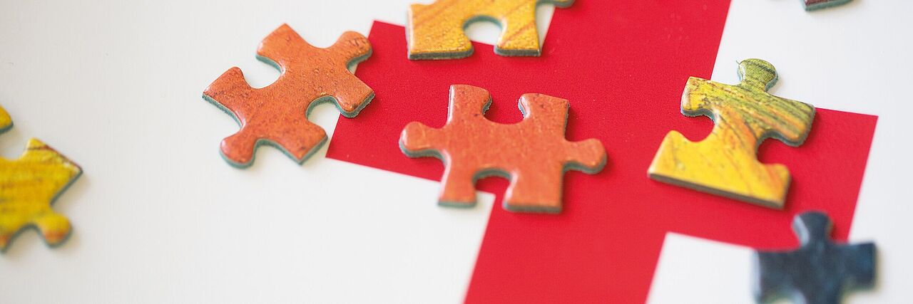 Ein rotes Kreuz ist auf einer weißen Fläche gedruckt. Auf dem Kreuz liegen viele kleine Puzzleteile. Sie sollen das komplexe Zusammenwirken des DRK symbolisieren.