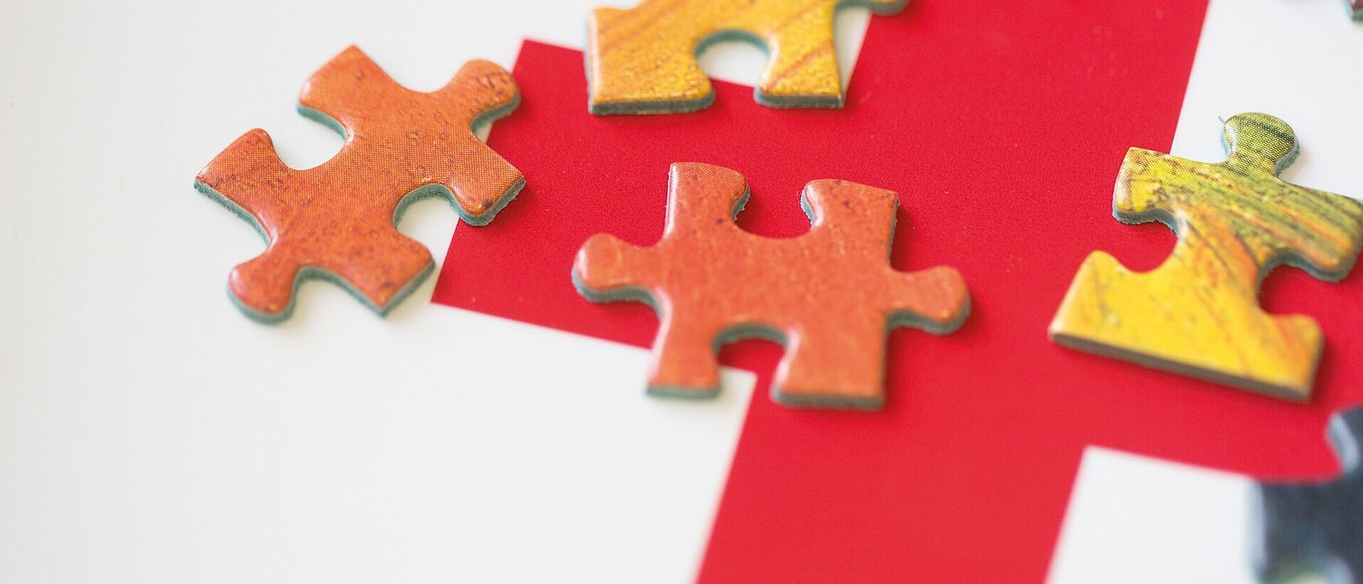 Ein rotes Kreuz ist auf einer weißen Fläche gedruckt. Auf dem Kreuz liegen viele kleine Puzzleteile. Sie sollen das komplexe Zusammenwirken des DRK symbolisieren.