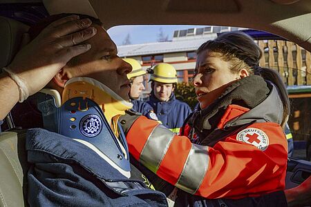 Eine Notfallsanitäterin lehnt sich nach innen, durch die offene Fahrertür eines Wagens. Sie leistet Erste Hilfe bei einem Verletzten auf dem Fahrersitz. Es handelt sich um eine Übung.